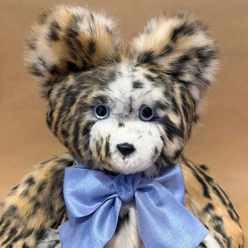 A plush leopard-style teddy bear with tan, black, and cream spotted fur, blue eyes, and a large light blue satin bow around its neck, photographed against a plain tan background.