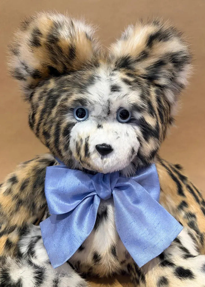 A plush leopard-style teddy bear with tan, black, and cream spotted fur, blue eyes, and a large light blue satin bow around its neck, photographed against a plain tan background.
