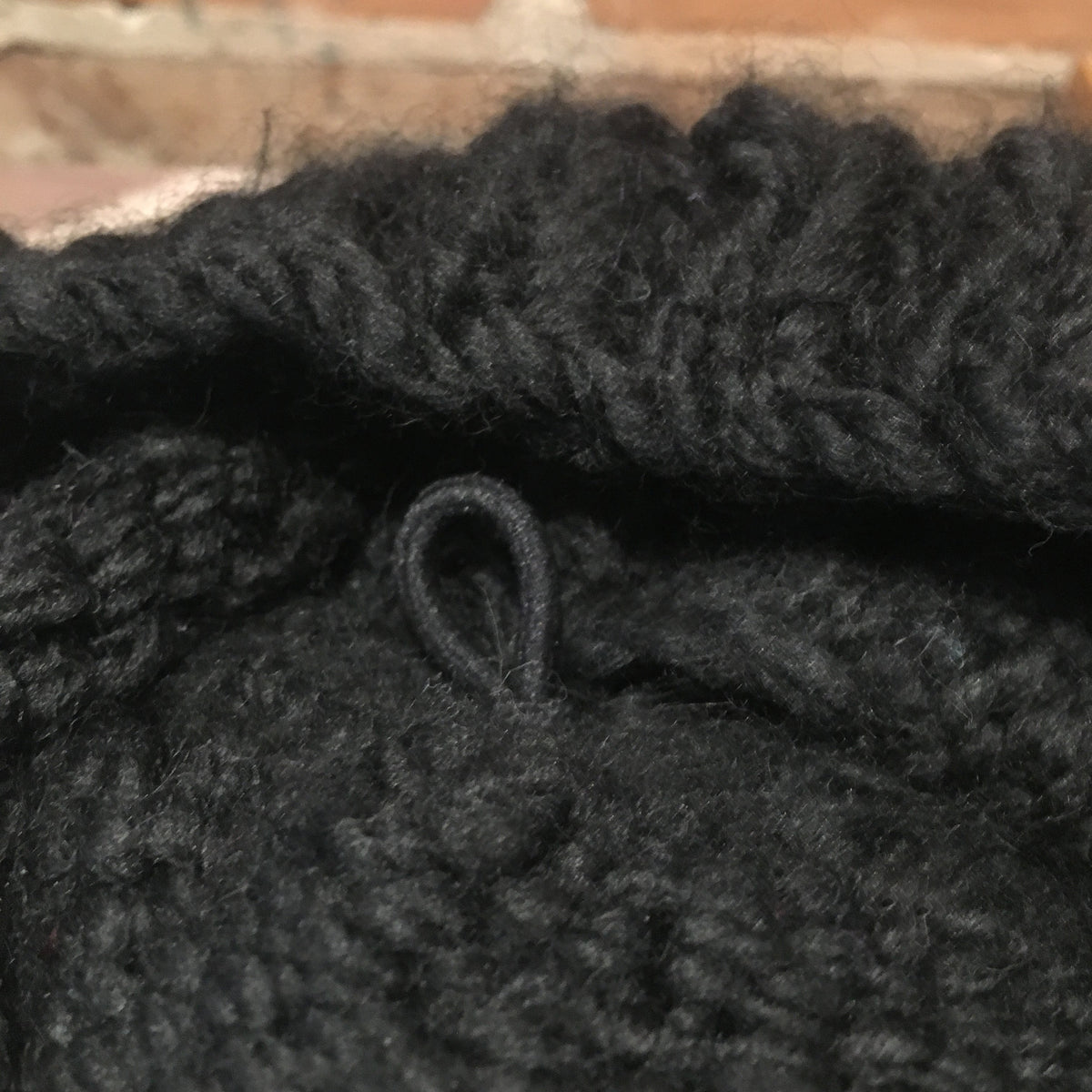 Close-up of a chunky black knitted fabric with a small black loop detail, set against a blurred brick background.