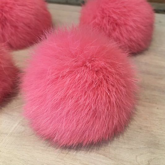 A close-up of a bright pink fluffy rabbit fur pom-pom resting on a light wood surface, with additional pom-poms partially visible in the background.