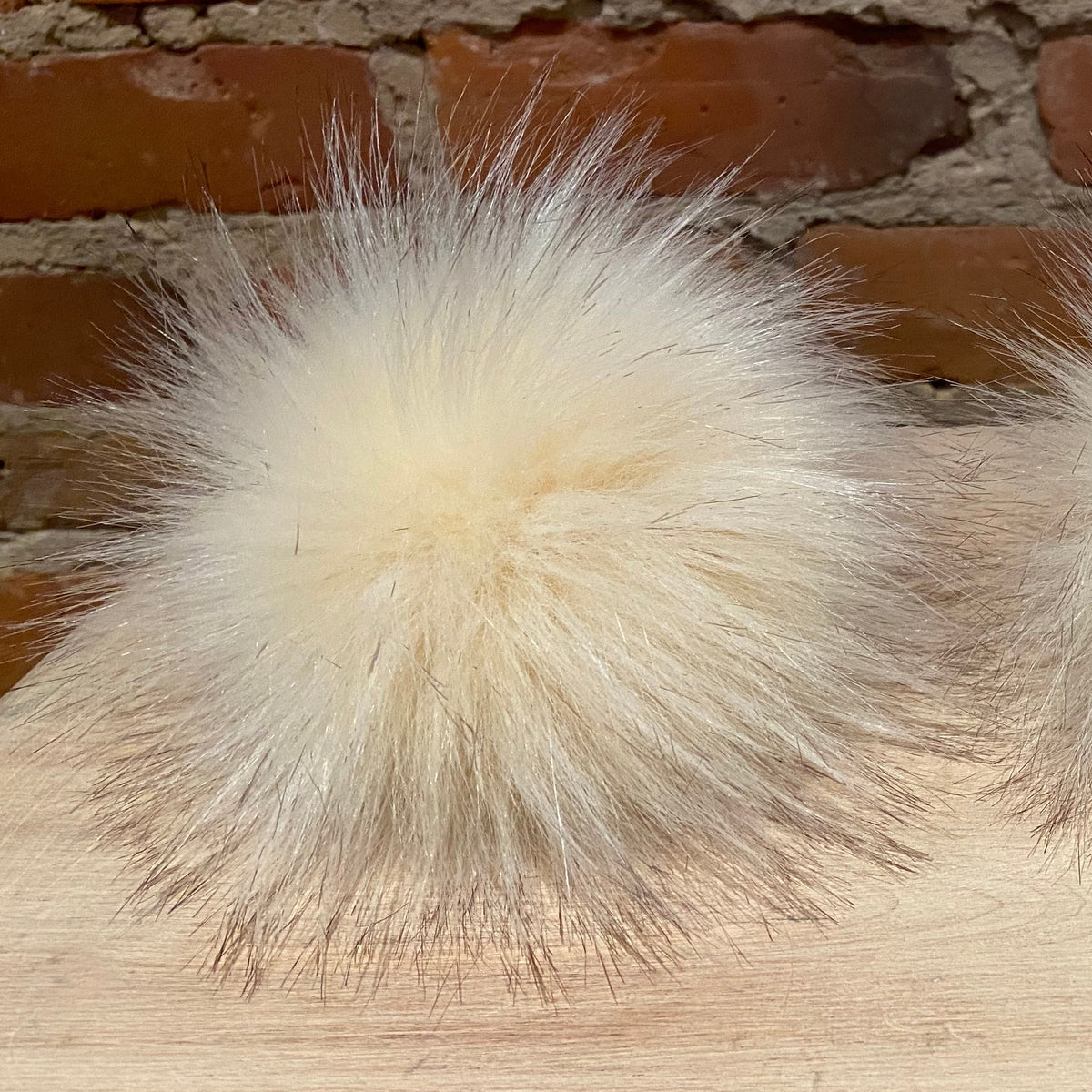 A fluffy cream-colored faux fur pom-pom accessory resting on a light wooden surface with a rustic brick wall background.