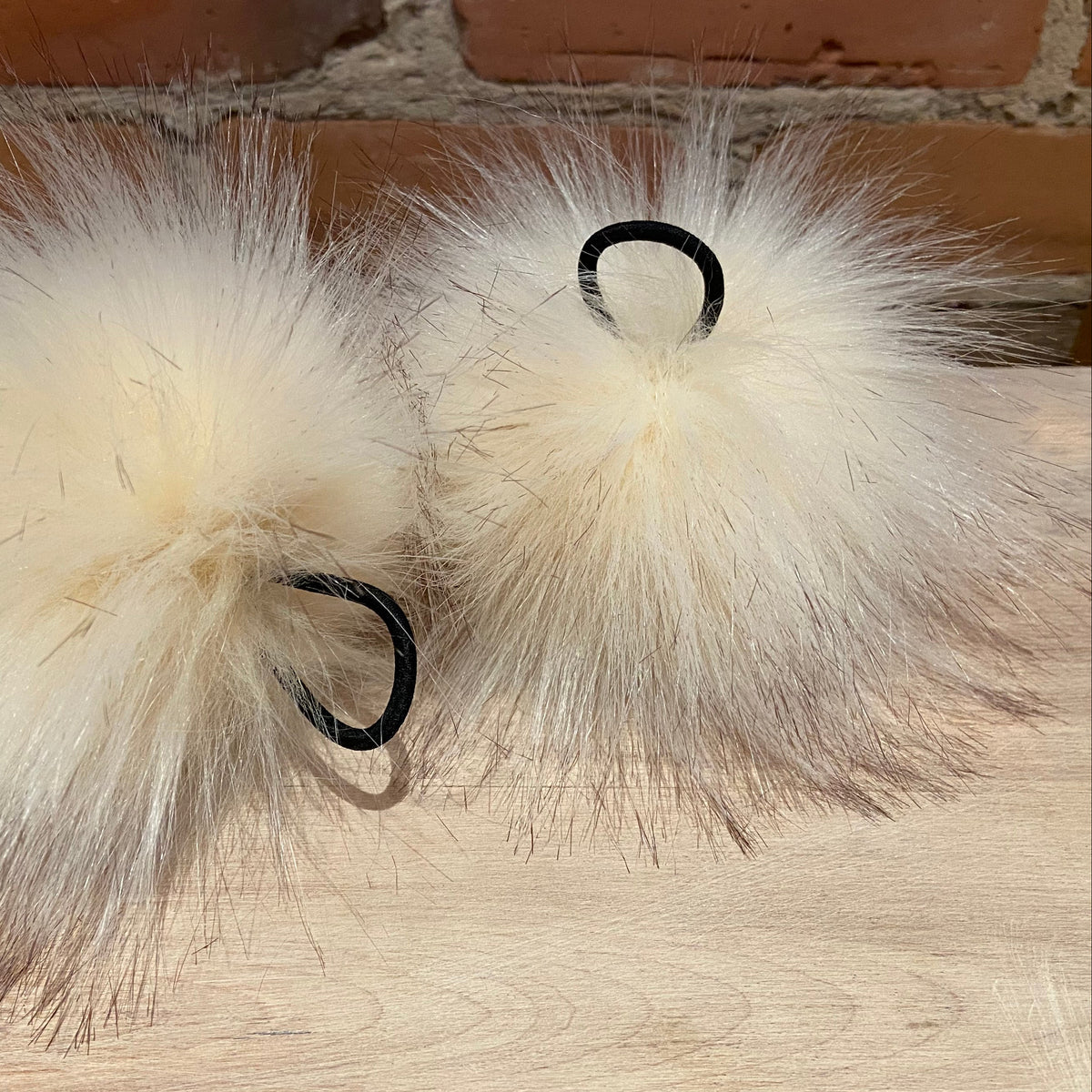Two fluffy cream faux-fur pom-pom hair accessories with black elastic loops, resting on a light wood surface with a brick wall in the background.