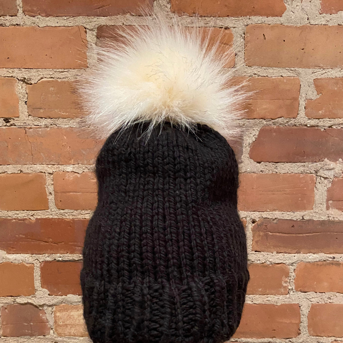 A black ribbed knit beanie hat with a large fluffy cream pom-pom, shown against a red-brown brick wall background.