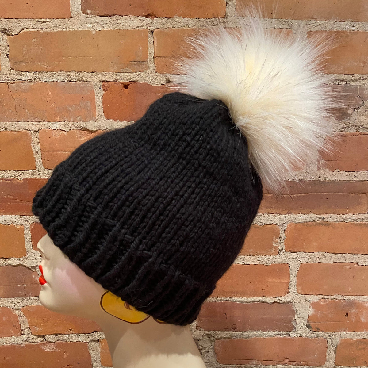A black ribbed knit beanie with a large fluffy cream faux-fur pom-pom, displayed on a mannequin head against a red-brown brick wall background.