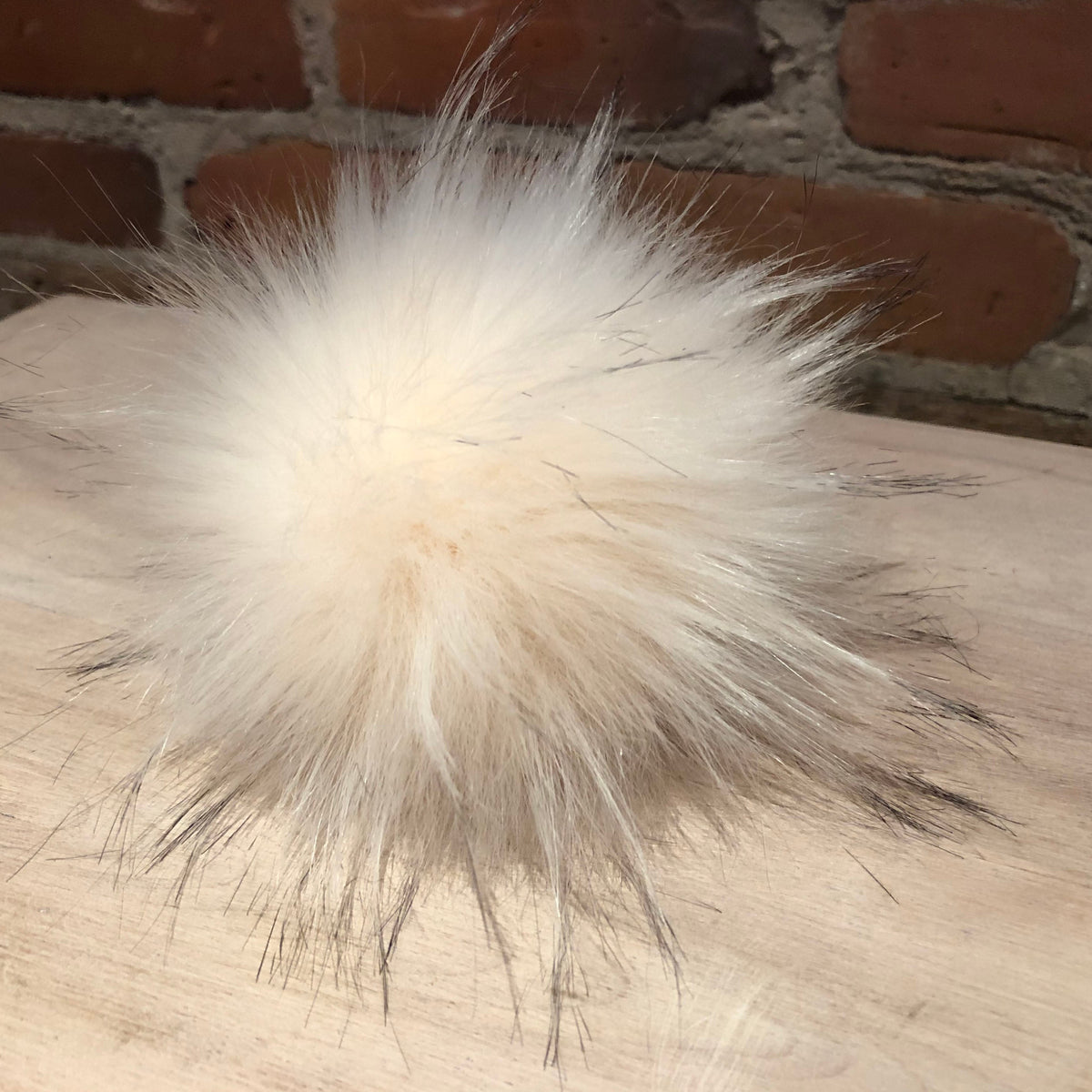 A fluffy faux fur pom-pom in cream and light brown, resting on a light wood surface with a brick wall background.