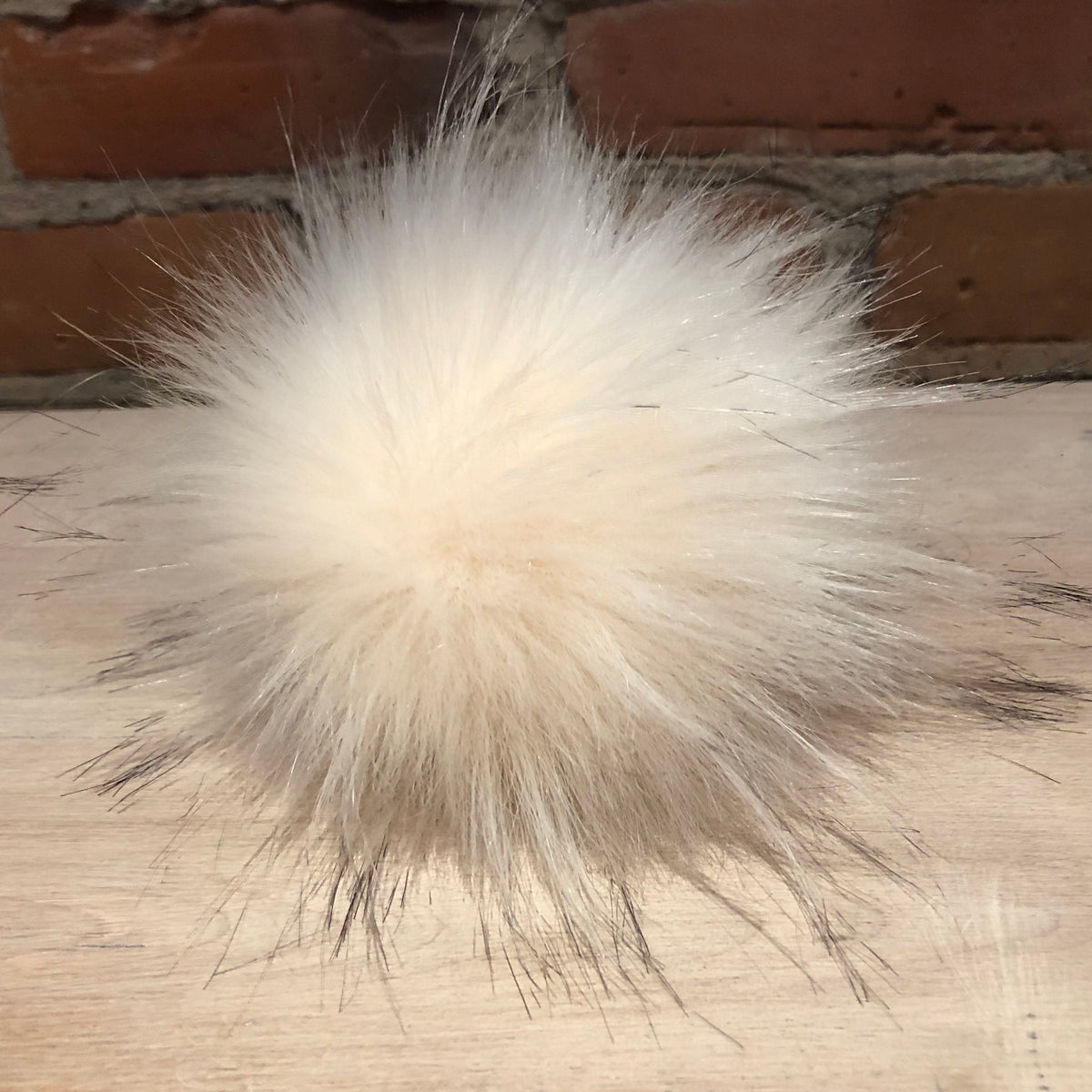 A fluffy cream-colored faux fur pom-pom with dark tips, resting on a light wood surface with a brick wall in the background.