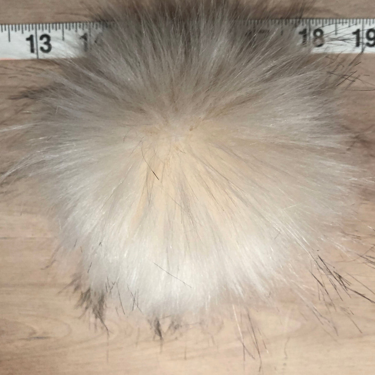 A fluffy faux fur pom-pom in a light beige and cream color, shown with a metal ruler at the top to indicate its size on a light wood background.