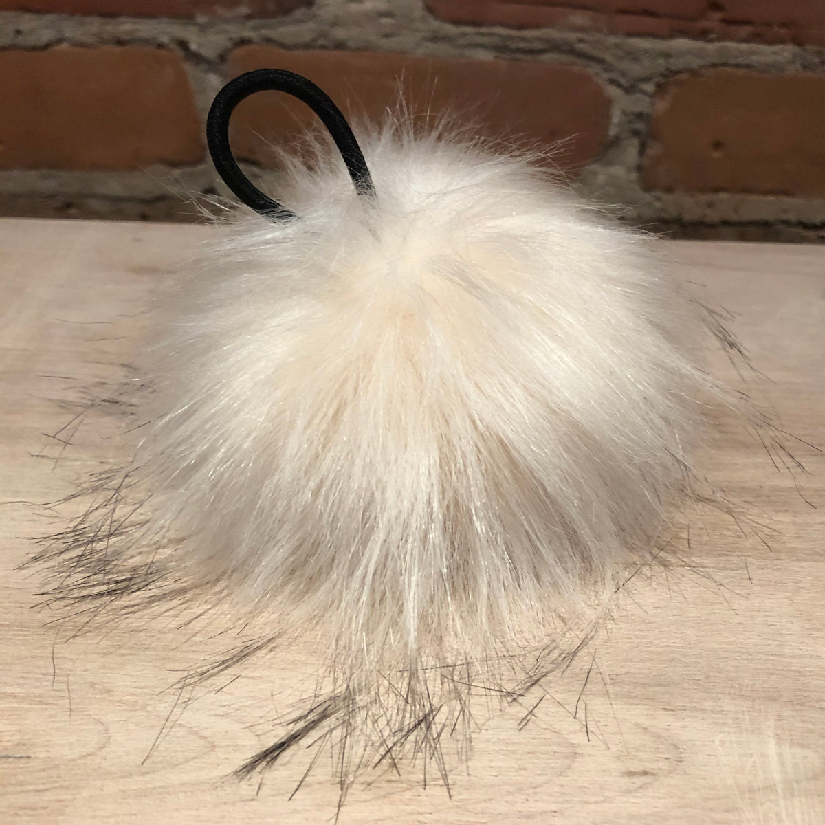 A fluffy cream faux fur pom-pom keychain or bag charm with a black elastic loop, resting on a light wood surface with a brick wall background.