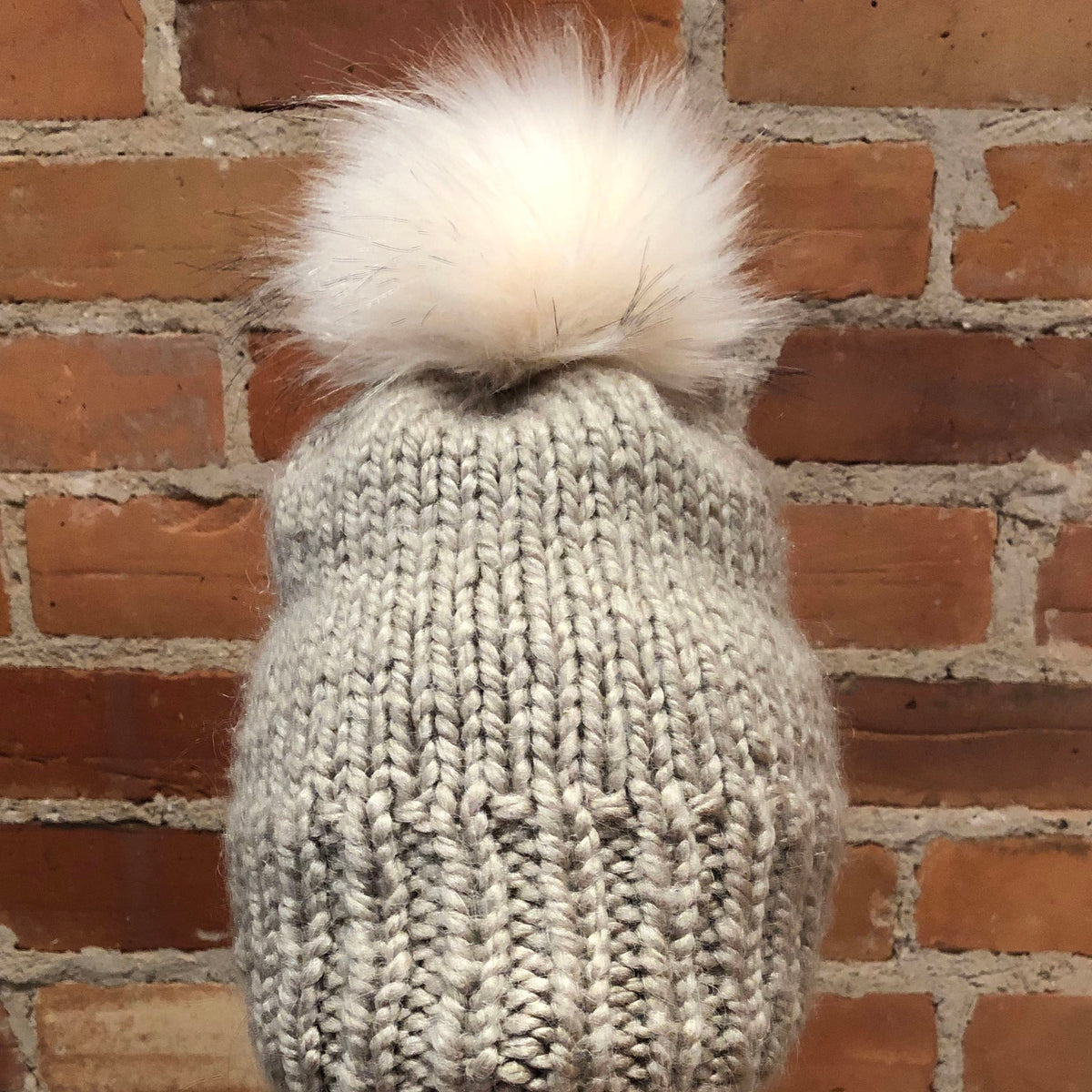 A light gray chunky knit beanie with a large fluffy cream pom-pom, shown against a rustic red-brown brick wall background.