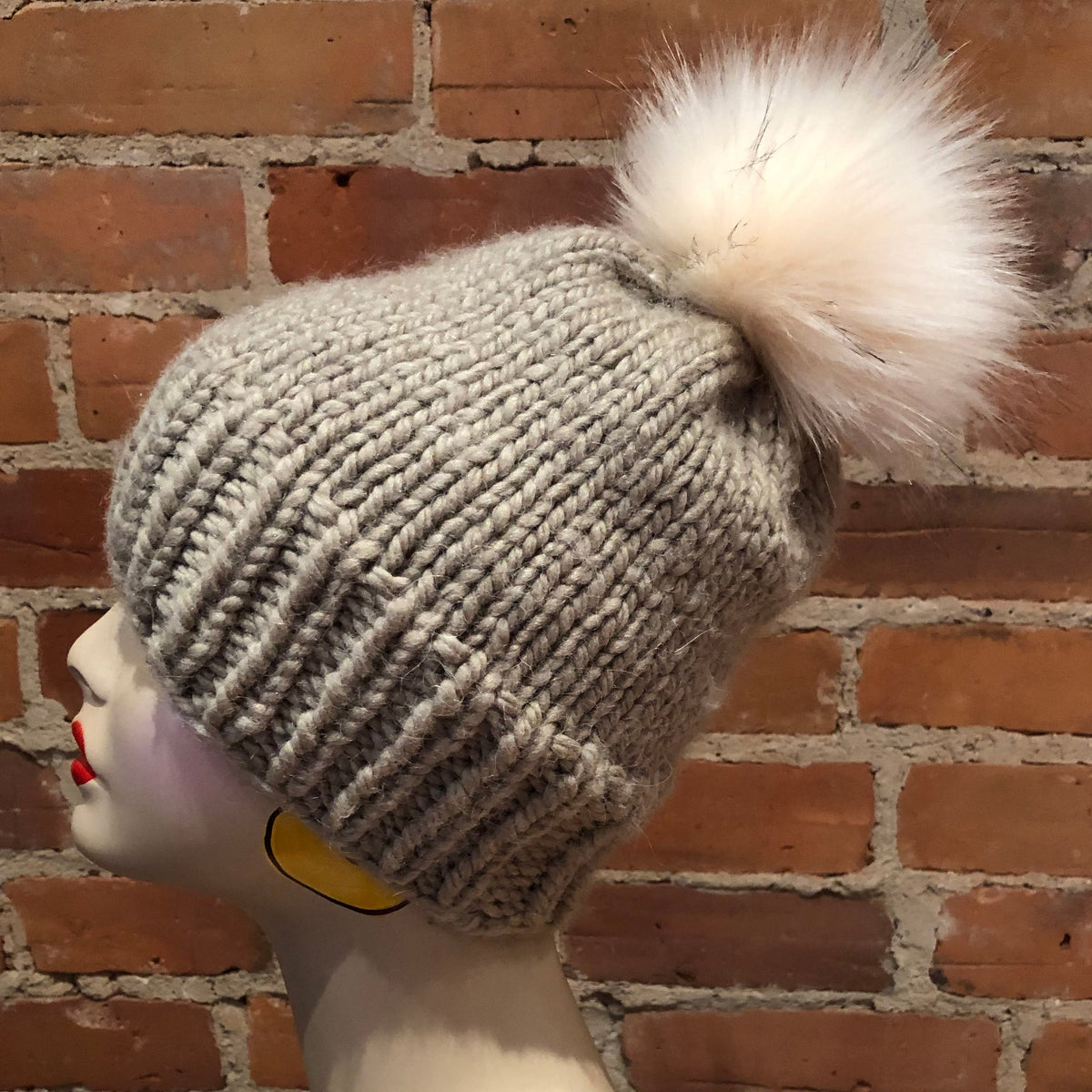 A light gray chunky knit beanie with a large fluffy cream faux-fur pom-pom, displayed on a mannequin head against a red brick wall background.