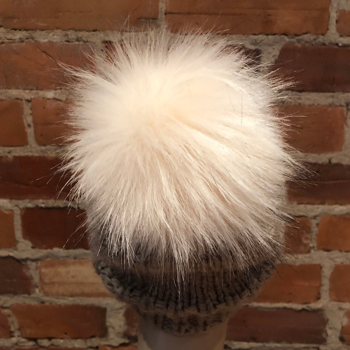 A fluffy cream-colored faux fur pom-pom hat displayed on a mannequin head against a red-brown brick wall background.