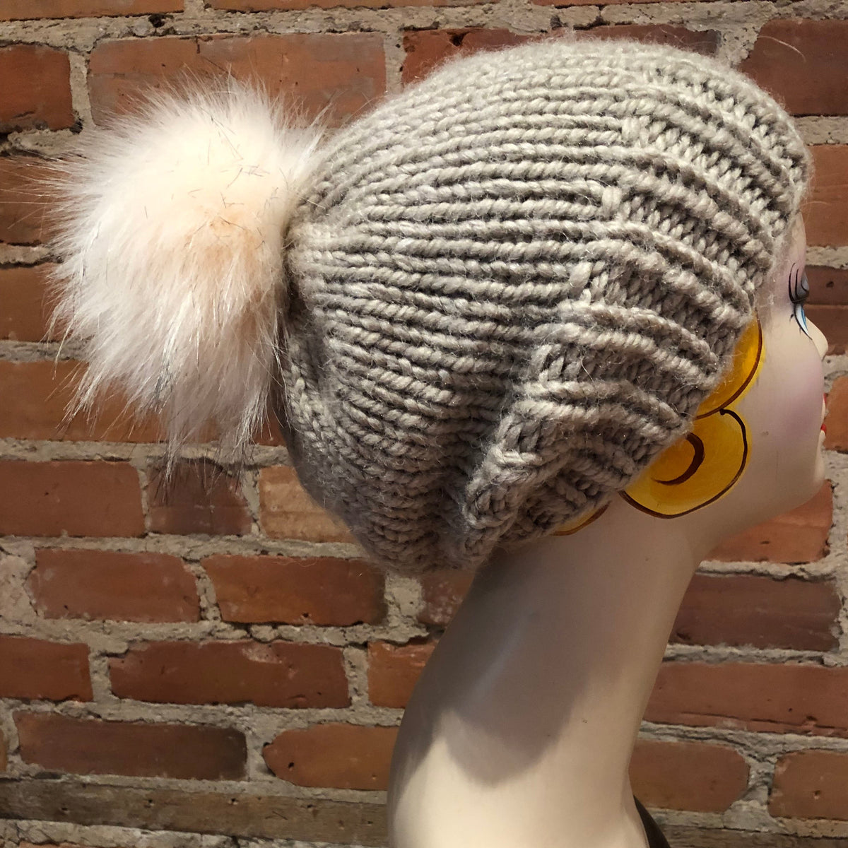 A light gray chunky knit beanie with a fluffy cream faux fur pom-pom, displayed on a mannequin head against a red brick wall background.