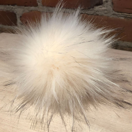 A large, fluffy, pale peach colored faux fur pom pom with black tip accents, displayed on a wooden surface with a brick wall in the background.