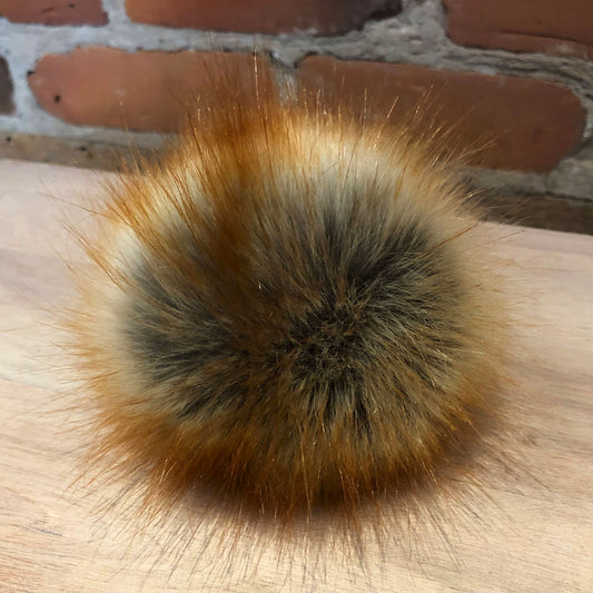 A round faux fur pompom in vibrant natural red and beige colors, with a dense bristly texture, displayed on a wooden surface with a brick wall in the background.